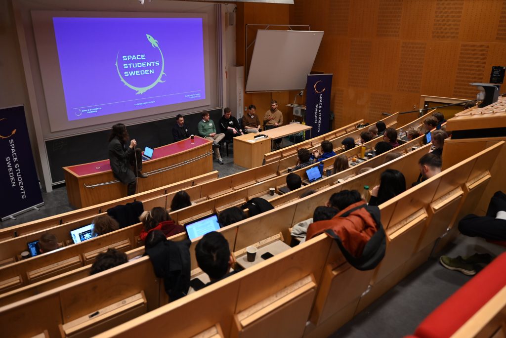 conf26_panel Wide-angle picture of participants in lecture hall. In the front there is a couple of people sitting on chairs. The Space Students Sweden logo is shown on the projector.